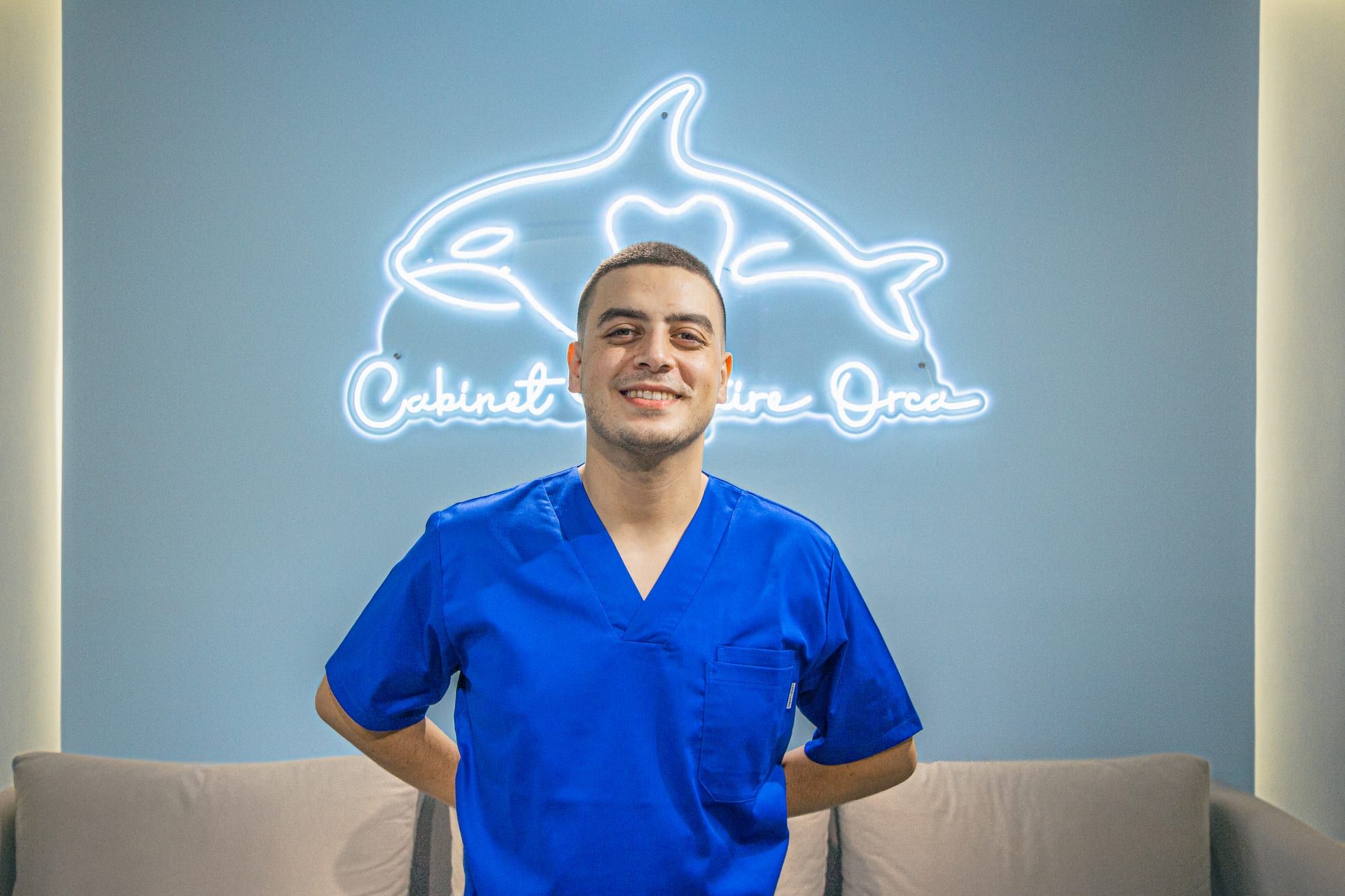 Dr Hamza Lahlou en tenue bleue, sous l’enseigne néon en forme d’orque du Cabinet Dentaire Orca
